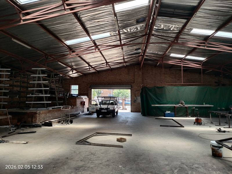 Spacious industrial warehouse featuring high-clearance steel truss roofing, skylights for natural illumination, exposed brick walls, and a large roller shutter door access.