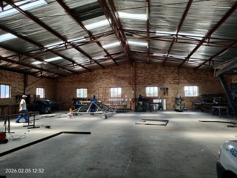 Spacious industrial workshop featuring exposed brick walls, high-pitched steel truss roof with skylights, and concrete flooring, suitable for manufacturing or storage operations.