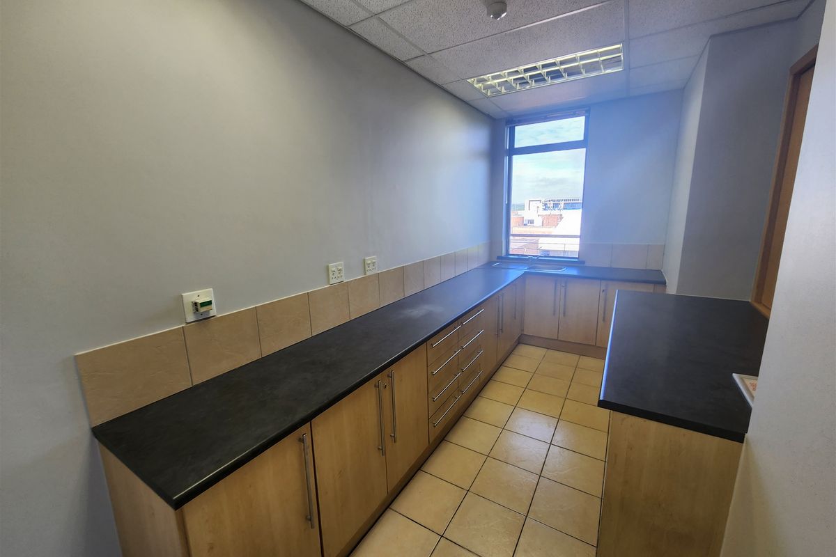 A functional office kitchenette featuring light wood cabinetry, dark laminate countertops, tiled flooring, and a drop ceiling with integrated lighting.