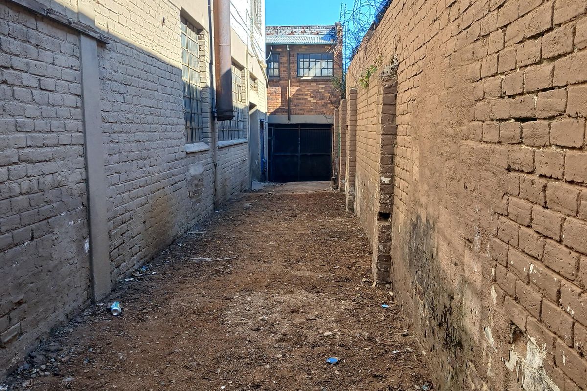 A narrow industrial alleyway featuring weathered brick boundary walls, razor wire security, and access to a rear metal roller shutter door.