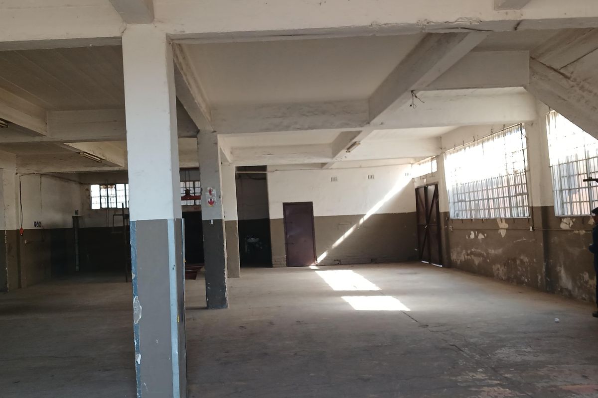 Spacious industrial utility area featuring exposed concrete beams, concrete flooring, and large windows with security bars, suitable for storage or workshop use.