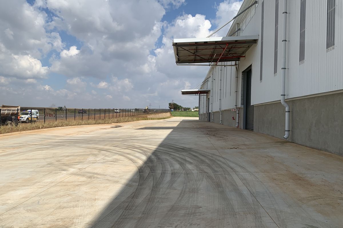 Exterior view of an industrial warehouse facility featuring a concrete loading yard, metal cladding, and overhead canopy structures for loading access.