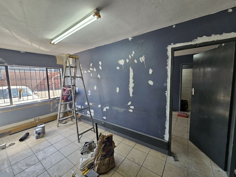 An industrial office space currently undergoing renovations, featuring tiled flooring, security bars on the window, and walls in the process of being repaired.
