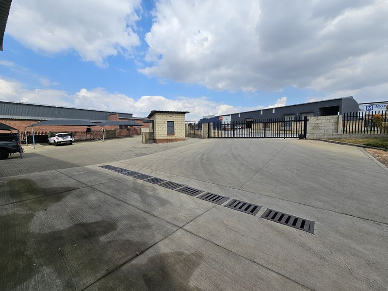 Industrial property entrance featuring a secure gated access point, guard house, paved driveway with drainage, and covered parking bays.