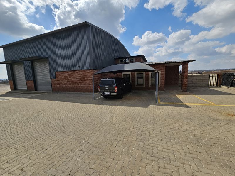 Industrial property featuring a large warehouse with roller shutter doors, brick facade, and a paved yard area with a shade cloth carport.