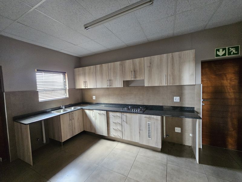 A functional industrial-style kitchen featuring light wood-finish cabinetry, dark laminate countertops, a gas hob, and tiled backsplash with neutral floor tiles.