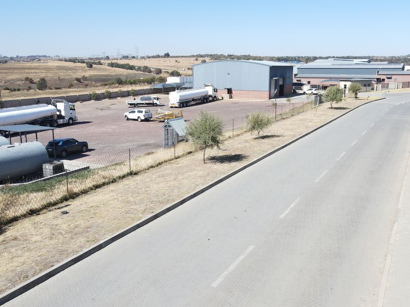 Aerial view of an industrial property featuring a large warehouse, paved yard area, storage tanks, and perimeter fencing with direct road access.