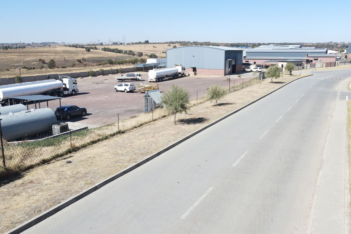 Aerial view of an industrial property featuring a large warehouse, paved yard area, storage tanks, and perimeter fencing with direct road access.