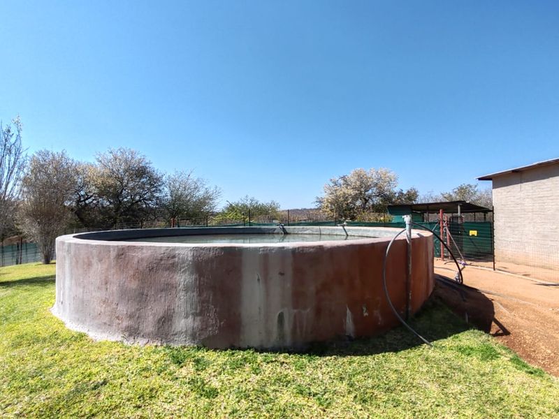 A circular concrete water reservoir situated in a garden setting, featuring basic plumbing connections and perimeter fencing.