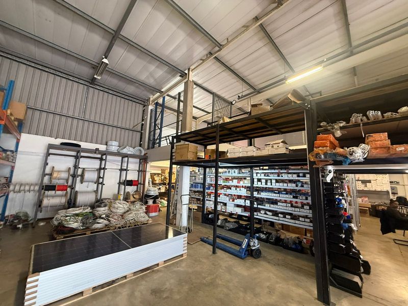 Industrial warehouse space featuring high ceilings, exposed steel structural elements, a mezzanine level, and extensive built-in heavy-duty shelving for storage.