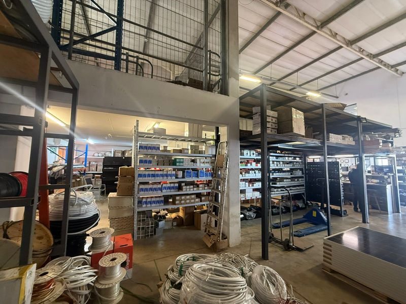 Industrial warehouse space featuring high ceilings with exposed steel trusses, a mezzanine level with security mesh, and extensive built-in metal racking systems.