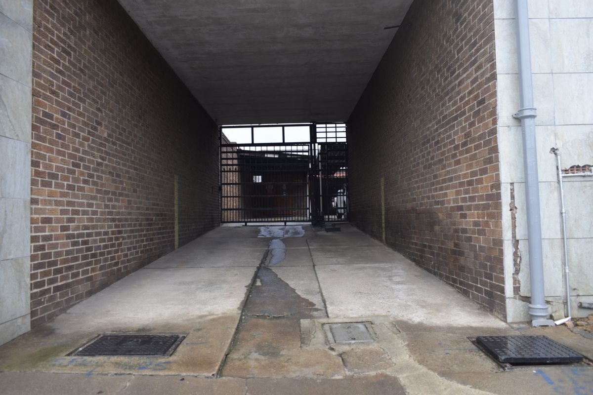 A covered rear access passage featuring brick walls, a concrete driveway with drainage, and a secure metal gate at the end.