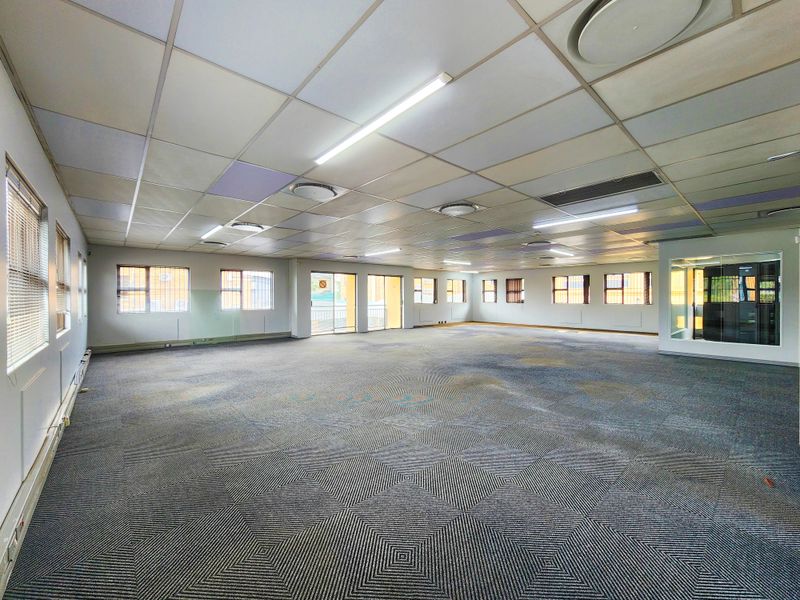 Spacious open-plan office area featuring suspended ceilings, fluorescent lighting, and carpeted flooring with large windows providing natural light.