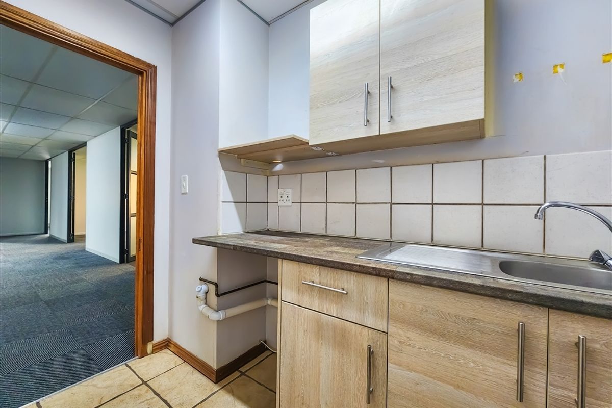 A functional office kitchenette featuring light wood-finish cabinetry, a stainless steel sink, and tiled backsplash, adjacent to a carpeted office hallway.