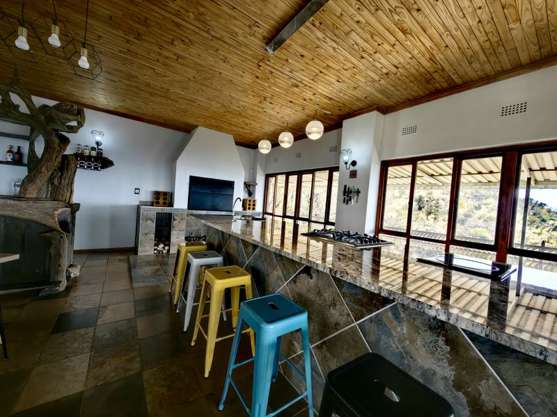 Spacious kitchen featuring a large granite breakfast bar, built-in braai, gas hob, and warm timber ceilings with expansive windows overlooking the landscape.