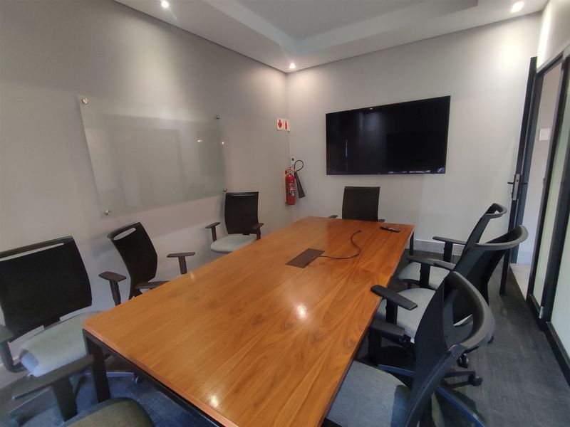 A professional office meeting room featuring a large wooden conference table, wall-mounted display screen, glass whiteboard, and recessed ceiling lighting.