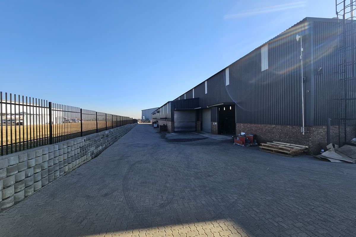 Exterior view of an industrial warehouse facility featuring a paved driveway, metal cladding, roller shutter doors, and a secure perimeter fence.