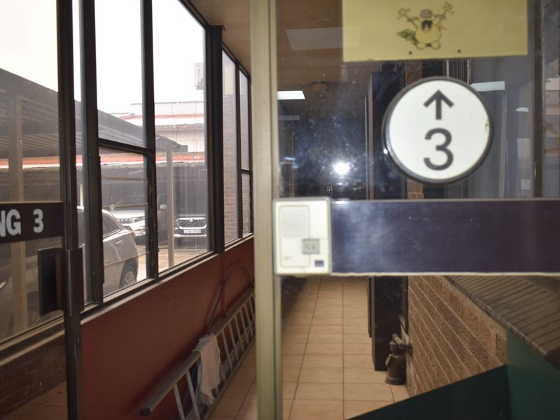 A functional office hallway featuring tiled flooring, brick wall accents, and large windows overlooking the parking area.