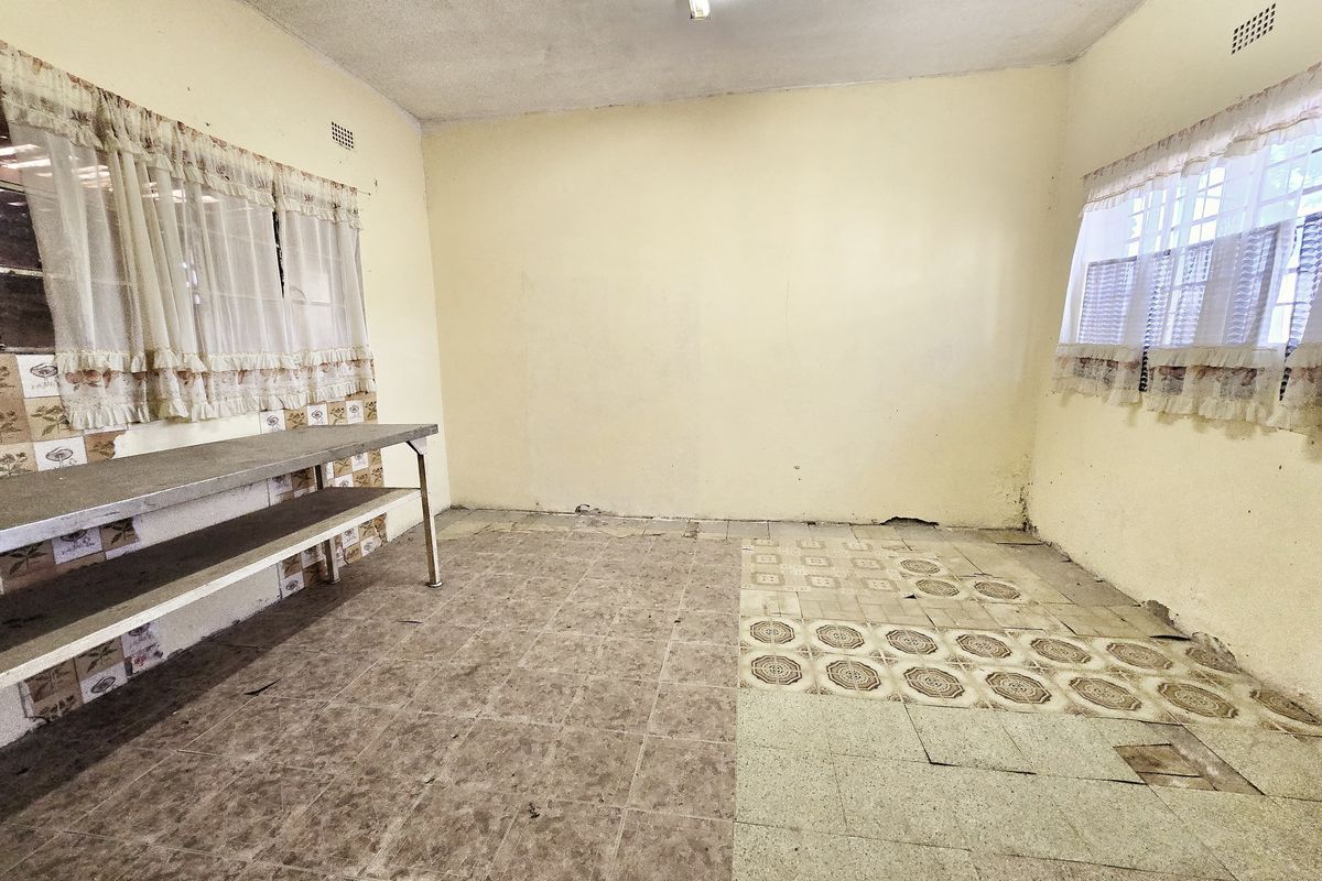 An empty, dated kitchen area featuring a stainless steel prep table, tiled splashbacks, and significant wear to the floor and wall finishes.
