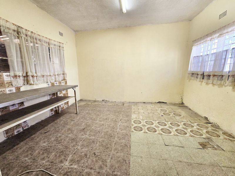 An empty, dated kitchen area featuring a stainless steel prep table, tiled splashbacks, and significant wear to the floor and wall finishes.