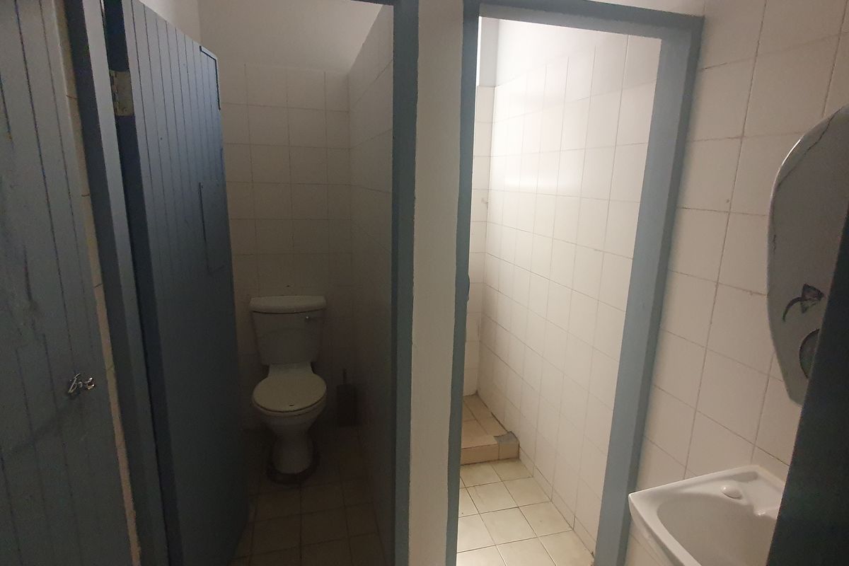 A functional commercial bathroom facility featuring tiled walls and floors, including a toilet cubicle and a small washbasin area.