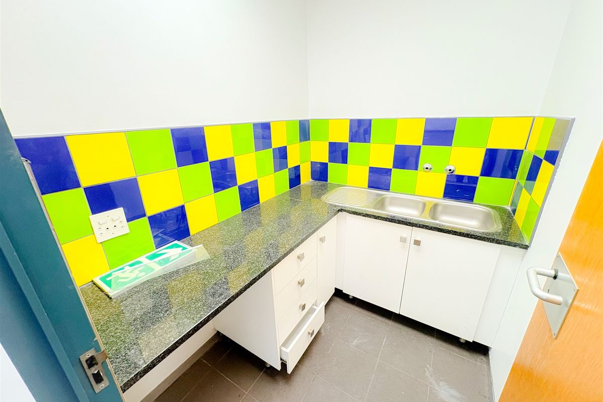 A compact office kitchenette featuring granite countertops, a double stainless steel sink, and vibrant multi-colored tiled splashbacks.