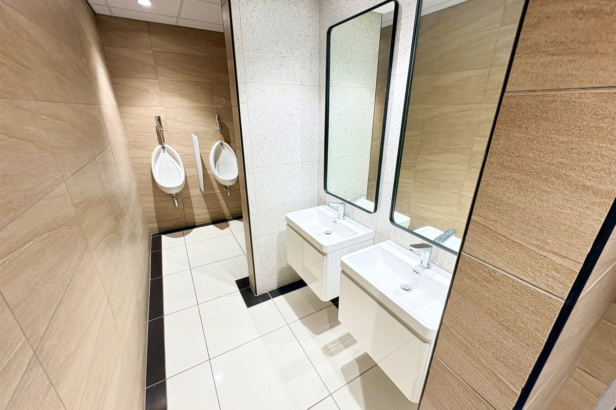 Modern office restroom featuring dual wall-mounted vanity basins with mirrors and two wall-hung urinals, finished with contemporary neutral tiling.