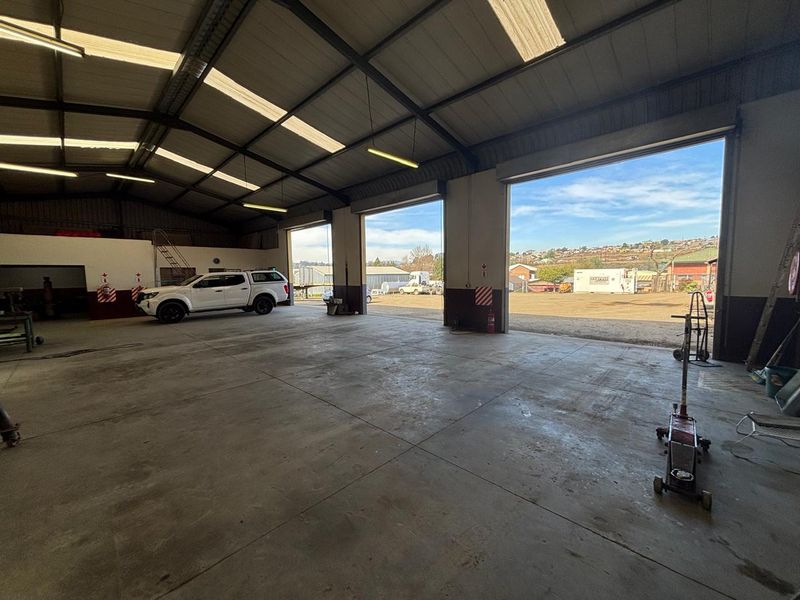Spacious industrial warehouse featuring high ceilings, concrete flooring, multiple large roller shutter doors for vehicle access, and ample natural light through overhead skylights.