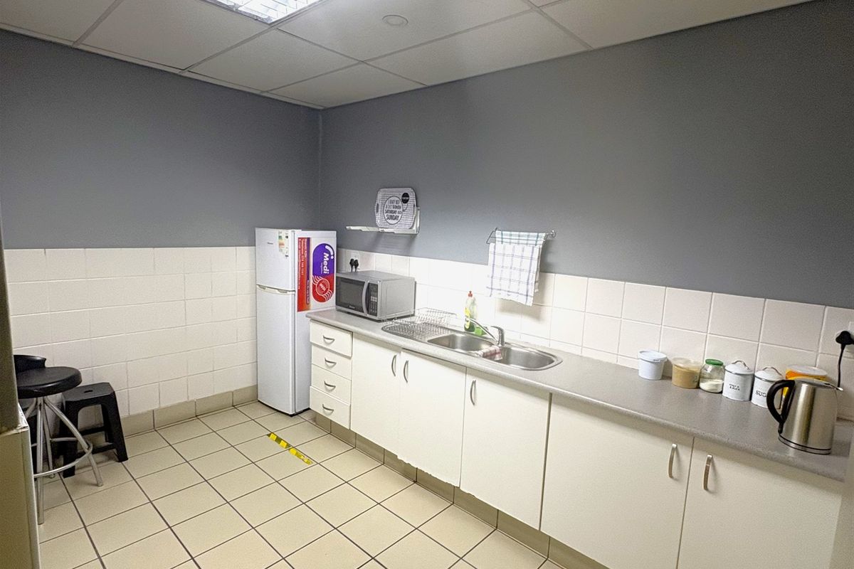 Functional industrial kitchenette featuring built-in cabinetry, stainless steel sink, tiled splashback, and durable ceramic flooring under a suspended ceiling.