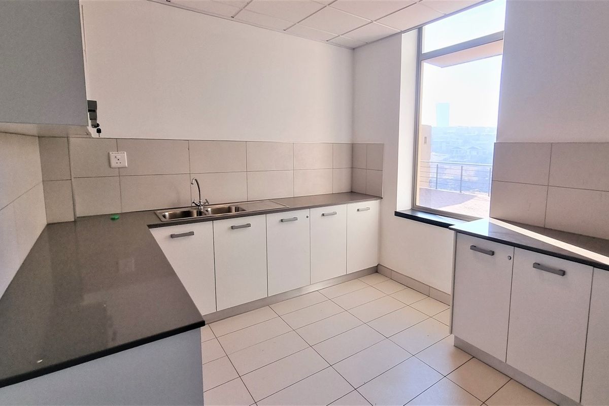 A clean, functional office kitchenette featuring white cabinetry, dark stone countertops, a stainless steel sink, and tiled flooring with natural light.