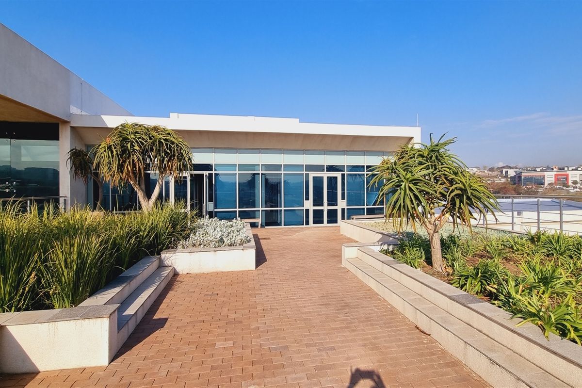 A modern office building exterior featuring a paved terrace, landscaped garden beds, and large glass curtain wall facade.