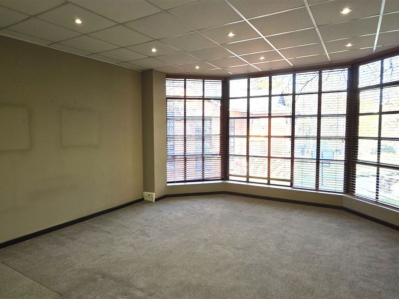 A spacious, empty office area featuring a large bay window with fitted blinds, recessed ceiling lighting, and neutral carpet flooring.