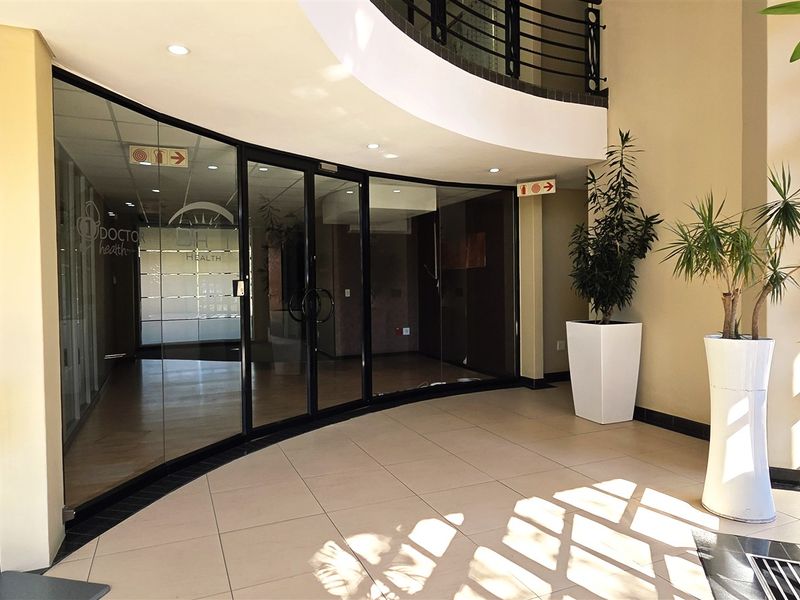 A bright and modern office building entrance featuring tiled floors, a curved glass partition, and a mezzanine level with a metal railing.