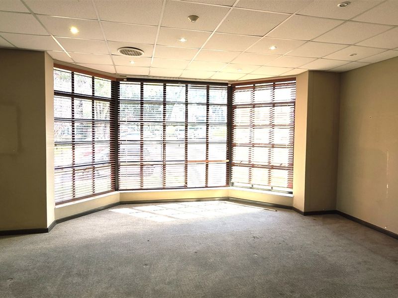 A vacant office space featuring a large bay window with fitted blinds, a suspended ceiling with recessed lighting, and carpeted flooring.