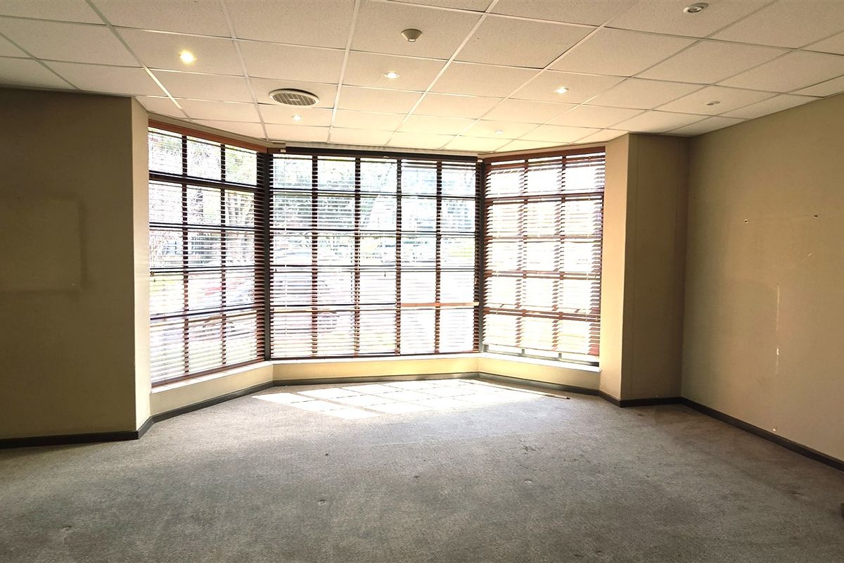 A vacant office space featuring a large bay window with fitted blinds, a suspended ceiling with recessed lighting, and carpeted flooring.