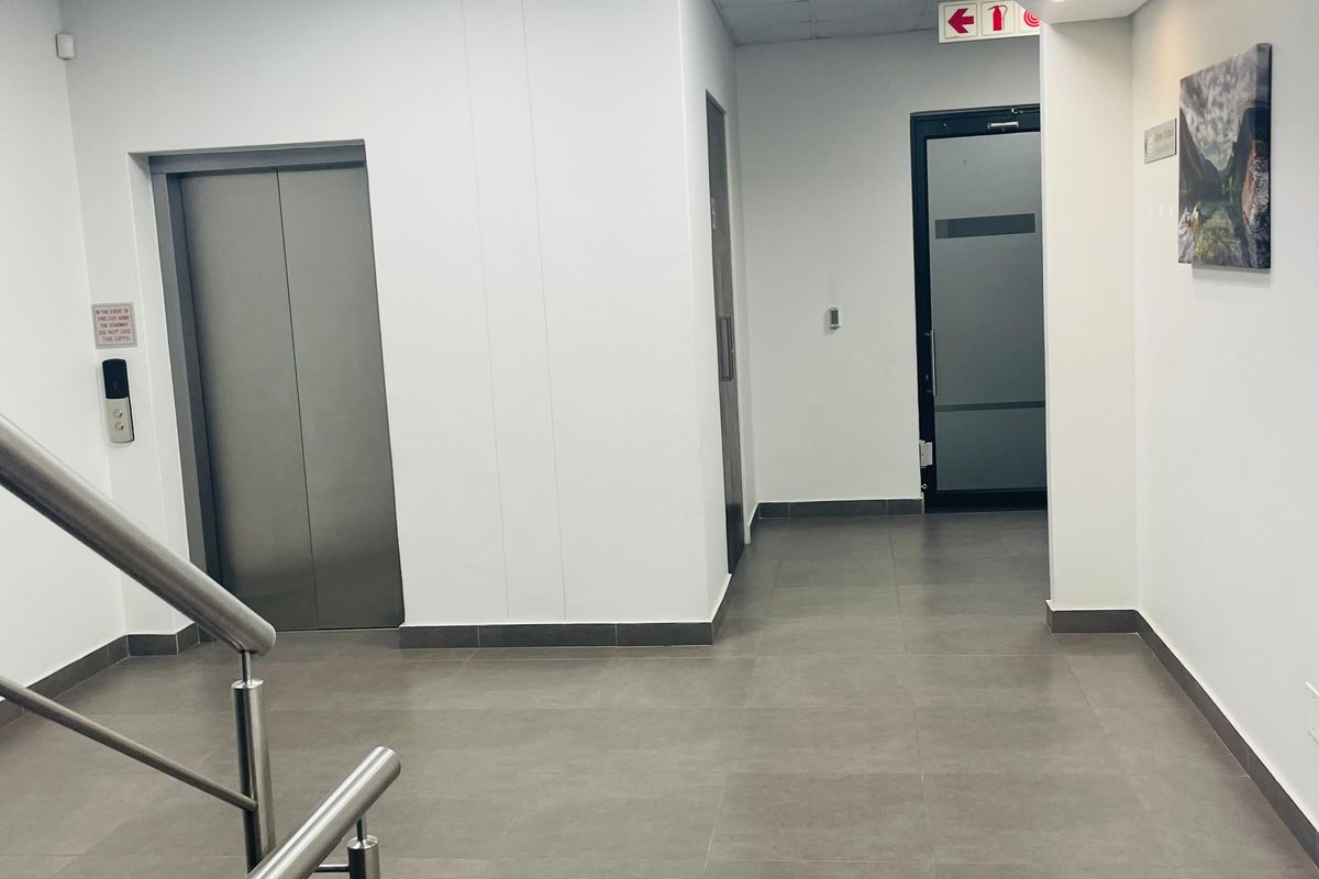 A clean, modern office building hallway featuring tiled floors, a stainless steel elevator, and professional recessed lighting.