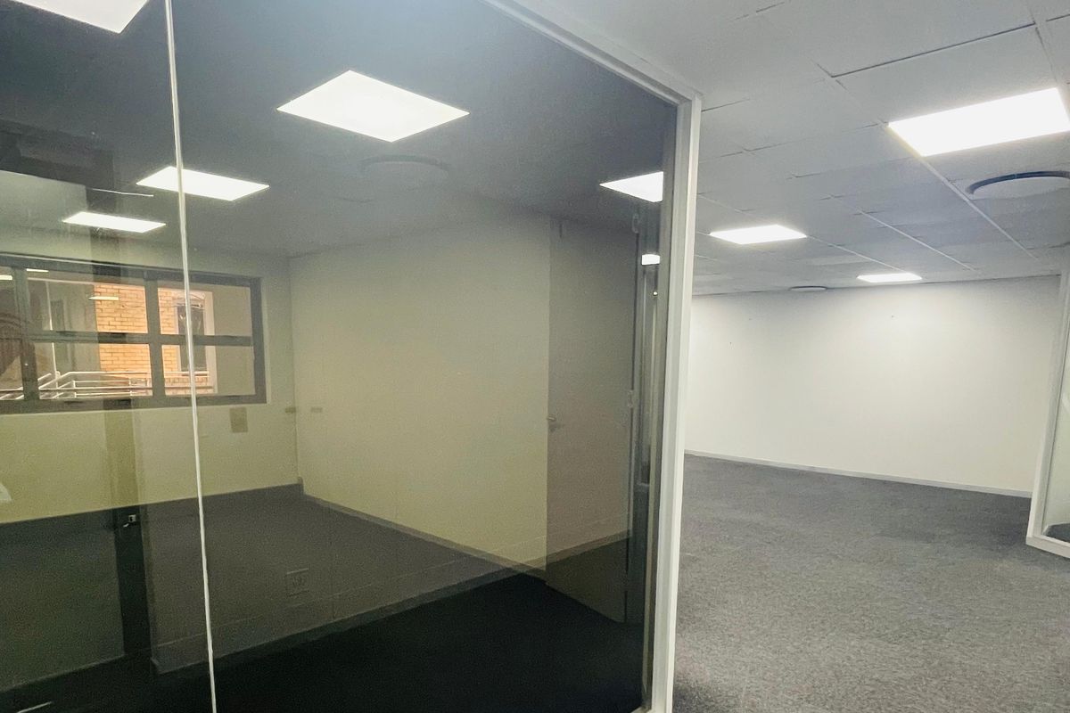 A spacious, empty office area featuring modern glass partitioning, suspended ceilings with recessed lighting, and neutral carpet flooring.