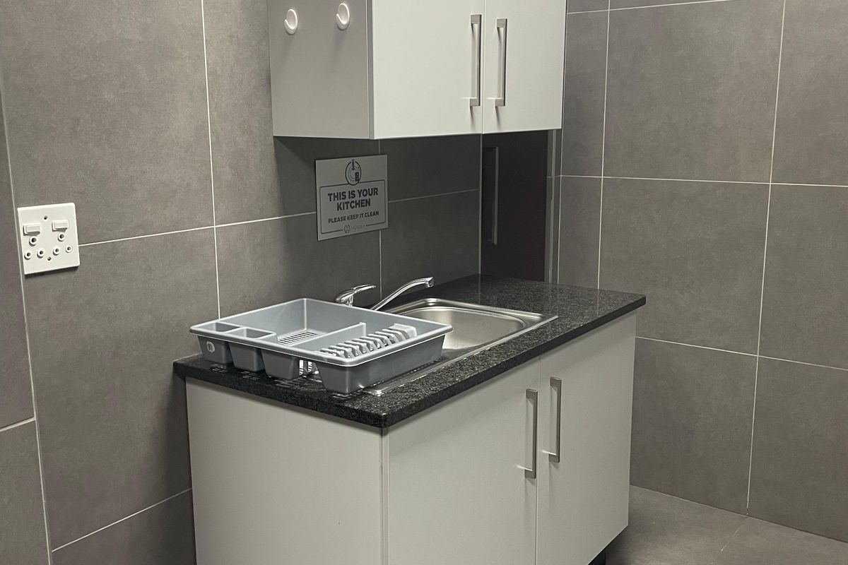 A compact office kitchenette featuring modern grey wall and floor tiles, white cabinetry, and a granite countertop with a stainless steel sink.