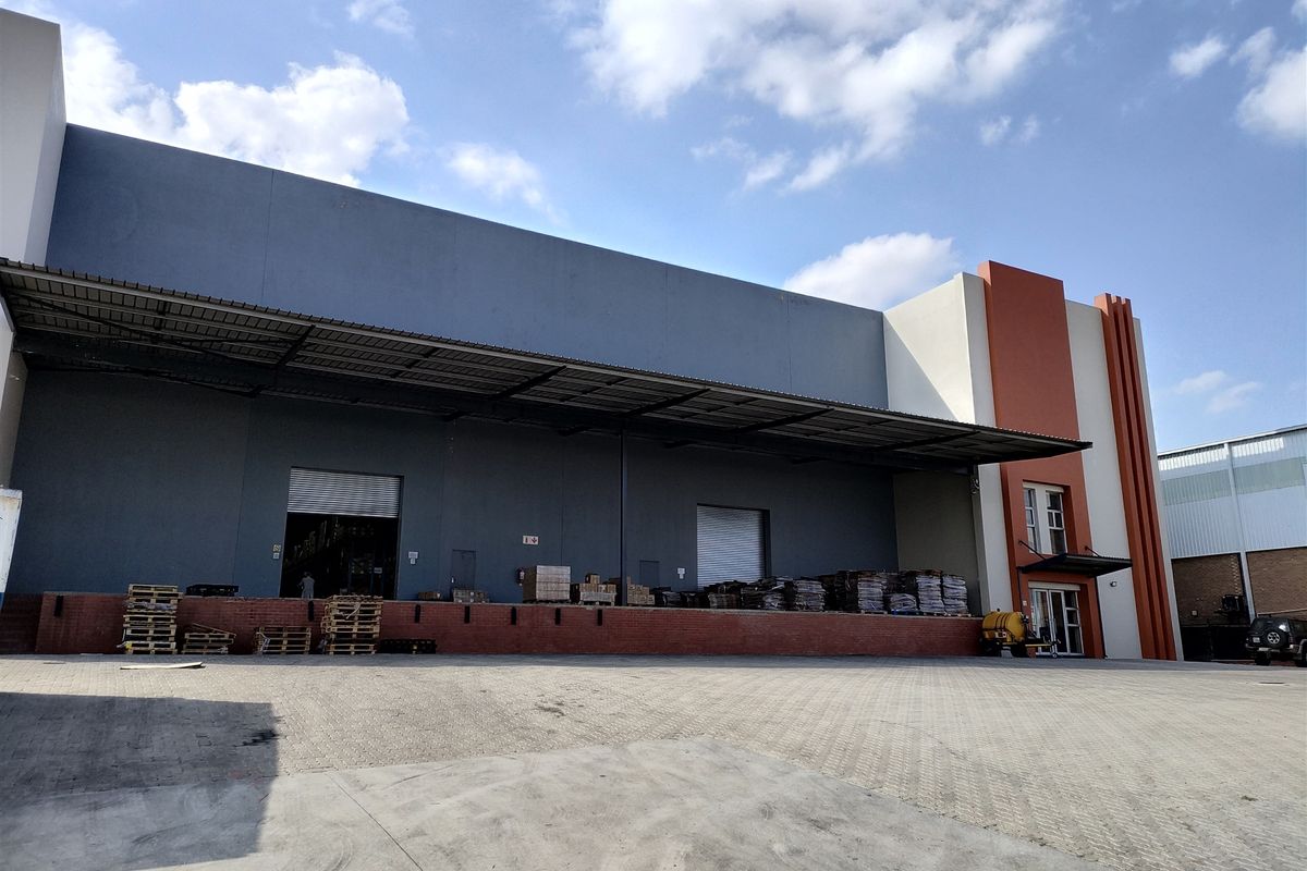 Industrial warehouse exterior featuring a large paved loading yard, multiple roller shutter doors, and a protective metal canopy over the loading bay area.