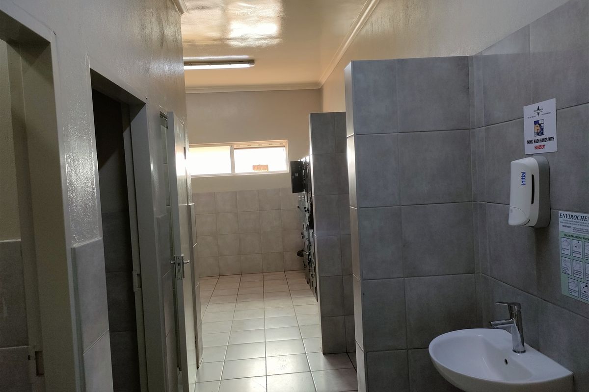 A functional industrial-style communal bathroom featuring tiled walls, tiled flooring, partitioned cubicles, and a pedestal washbasin.
