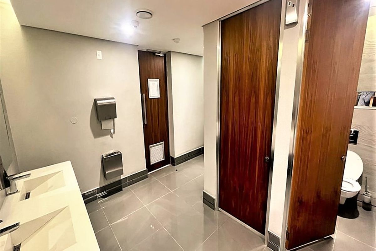Modern office restroom featuring dual vanity sinks, tiled flooring, and private wooden stall doors with integrated fixtures.