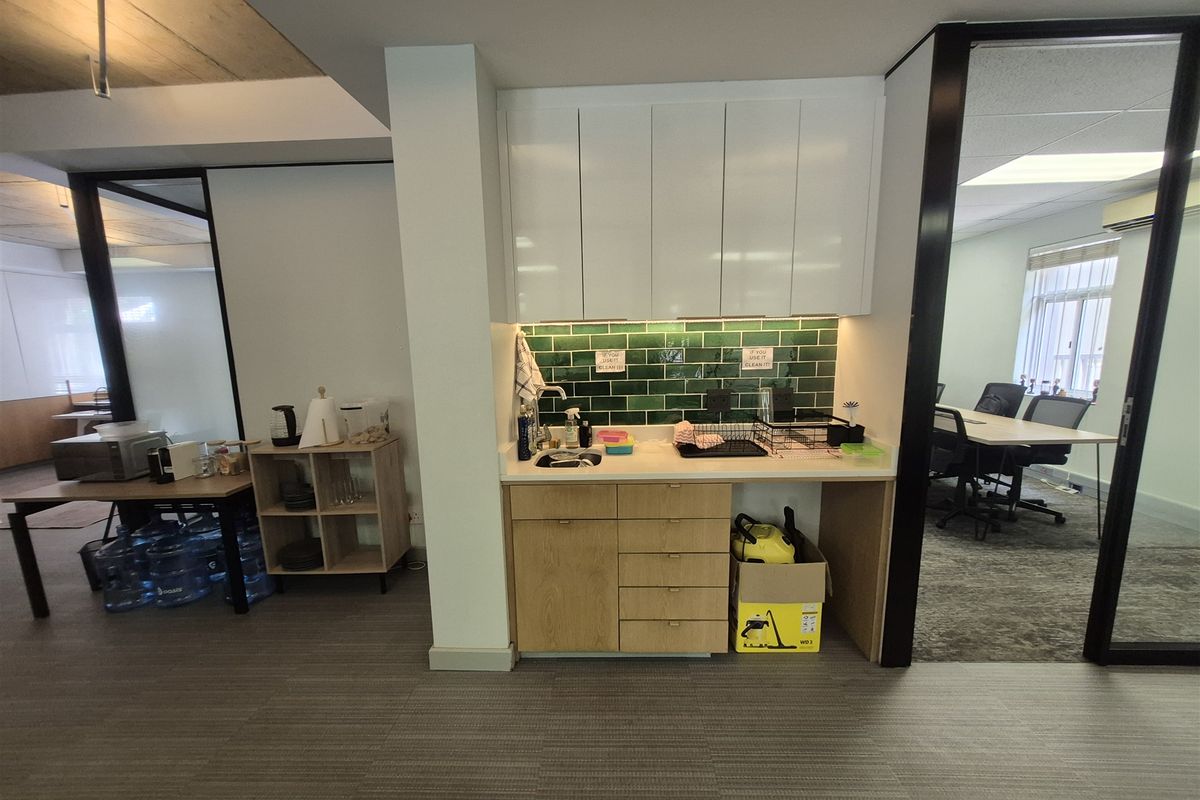 A compact office kitchenette featuring white upper cabinetry, light wood lower drawers, and a green subway tile backsplash with an integrated sink.