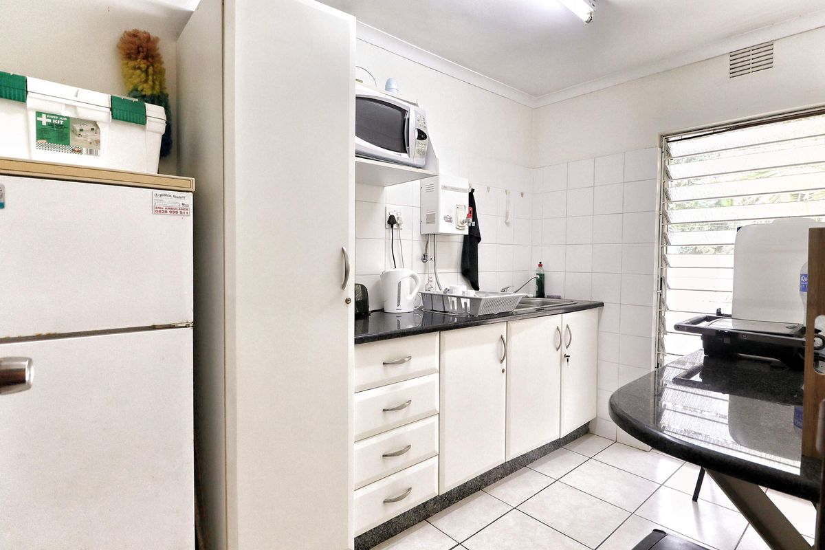 A functional office kitchenette featuring white cabinetry, granite countertops, tiled backsplash, and louvered windows for natural ventilation.