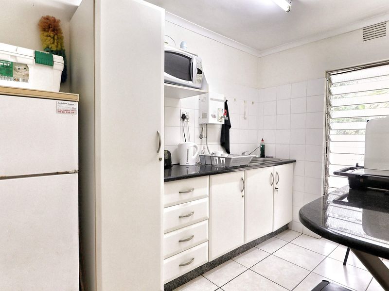 A functional office kitchenette featuring white cabinetry, granite countertops, tiled backsplash, and louvered windows for natural ventilation.