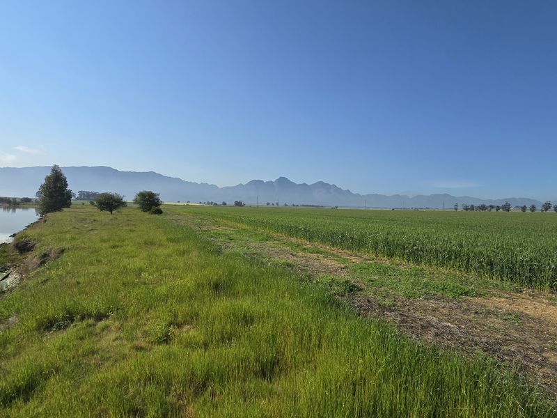 A scenic view of the farm property featuring a dam, expansive green fields, and distant mountain ranges under a clear blue sky.