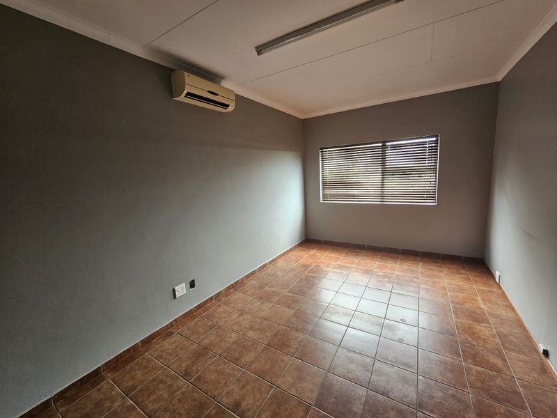 A clean, empty office space featuring tiled flooring, neutral grey walls, an integrated air conditioning unit, and window blinds for privacy.