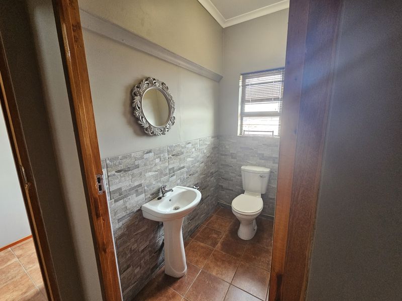 A functional office bathroom featuring a pedestal basin, toilet, tiled splashback, and tiled flooring with natural light from a window.