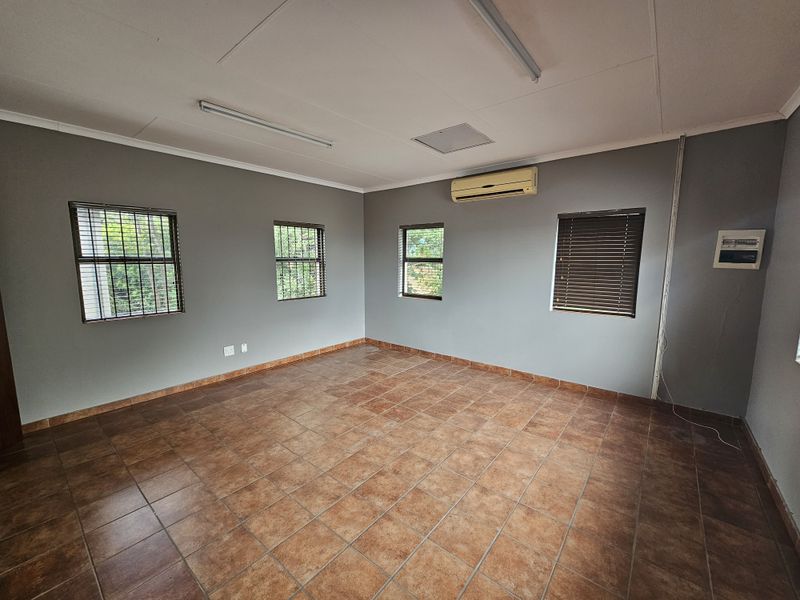 A spacious, empty office area featuring tiled flooring, multiple windows with security bars and blinds, and a wall-mounted air conditioning unit.