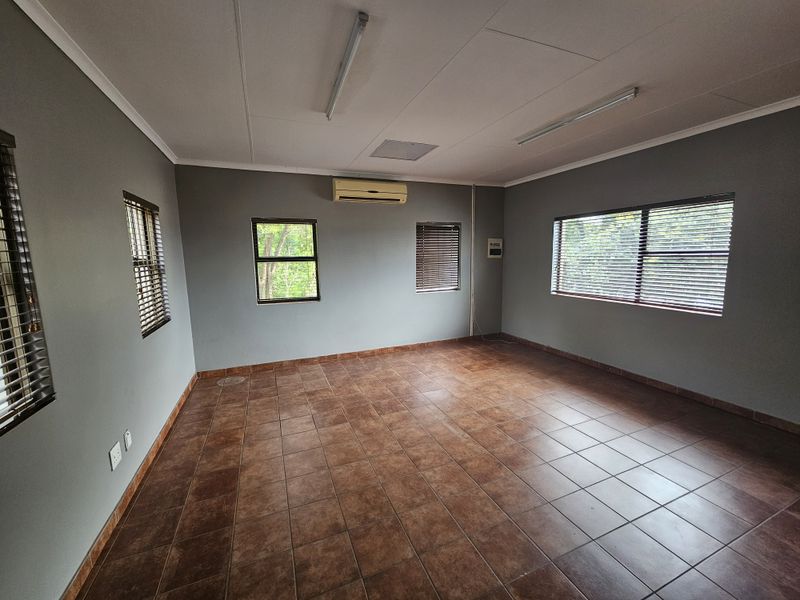 A spacious, empty office area featuring tiled flooring, neutral grey walls, fitted venetian blinds, and a wall-mounted air conditioning unit.