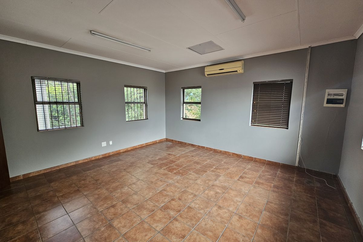 A spacious, empty office area featuring tiled flooring, multiple windows with security bars and blinds, and a wall-mounted air conditioning unit.
